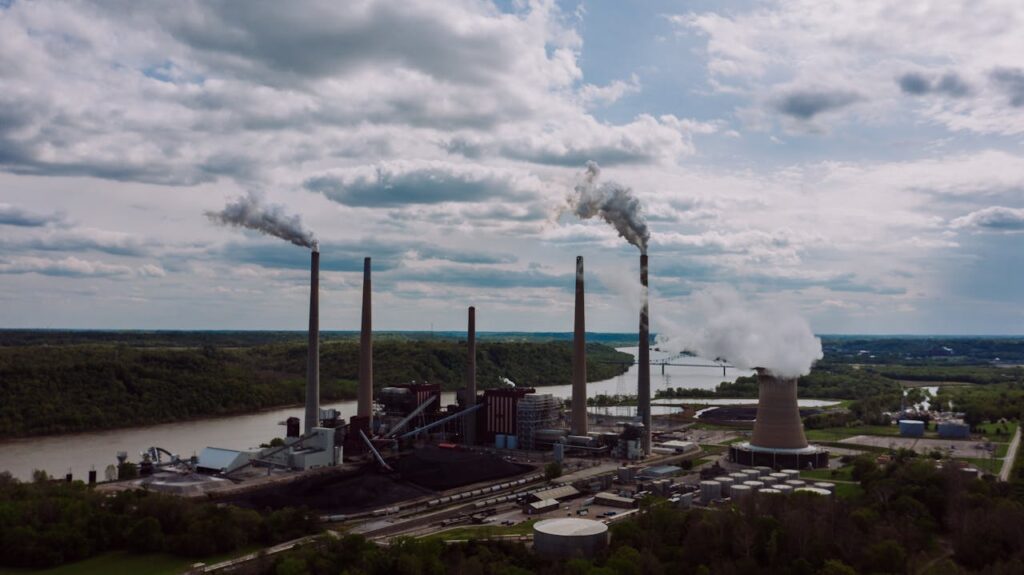 Air pollution by smoke coming out of factory chimneys located near river against cloudy sky on summer day